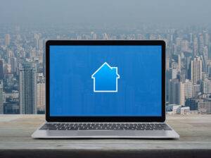 Haussymbol mit Kopierbereich auf modernem Laptop, auf Holztisch über Stadtturm und Wolkenkratzer, Geschäftsimmobilien-Online-Konzept