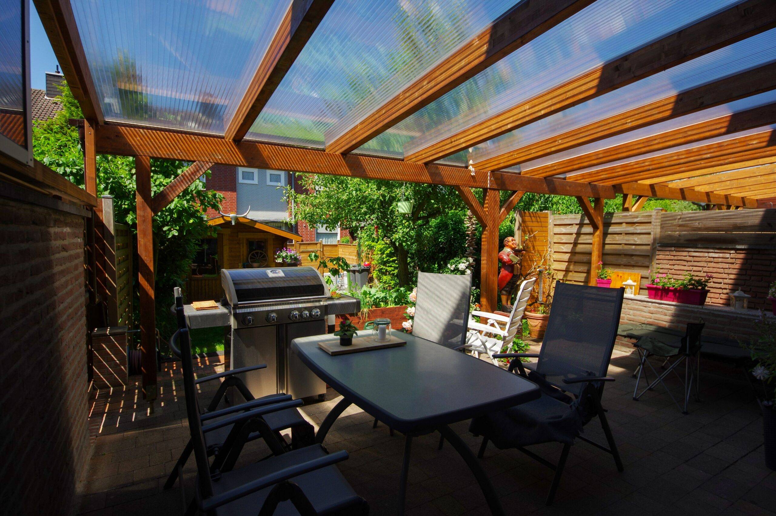 Terrassenüberdachung Köln aus Holz mit Gartenmöbeln und Grill unter klarer Überdachung im sonnigen Garten